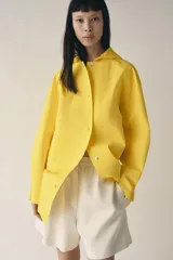 Cazadora impermeable de acabado engomado en color amarillo, con capucha ajustable, cierre frontal con botones a presión, bolsillos laterales y mangas largas ajustables.