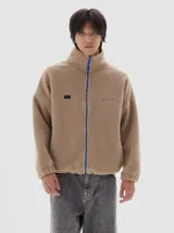 Campera de sherpa color beige con cuello alto, cierre frontal a contratono en color azul y bordado de la marca Pastiche en el pecho.