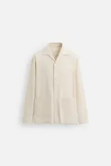 Sobrecamisa de hombre de corte relajado, color beige claro con finas rayas verticales en tono más oscuro. Presenta cuello bowling, manga larga con puño abotonado, bolsillos de parche en el pecho y en la cadera, y aberturas laterales en el bajo.