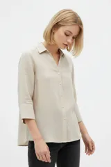 Camisa de algodón color beige, de corte holgado, con mangas tres cuartos, cuello camisero y cierre frontal de botones.