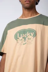 Remera de corte holgado con diseño color block, combinando paneles en color blanco y verde claro. Presenta un estampado frontal con la palabra Lucky dentro de un óvalo y motivos gráficos de flores.