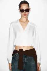 Camisa corta de tejido satinado color marrón, con cuello solapa, escote en pico y manga larga. Presenta cierre frontal con botones forrados a tono.