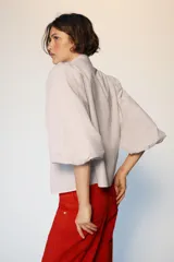 Blusa de cuello subido con escote en pico y cierre frontal con botones forrados. Presenta mangas abullonadas de largo tres cuartos con elástico en los puños y detalle de lazos en el cuello y el frente.