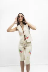 Chaleco tipo sastre color crema con estampado de rostros y flores, con escote en V y cierre frontal con lazos.