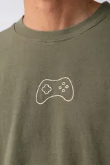 Remera de corte oversize color marrón, con cuello redondo y mangas cortas con hombros caídos. Presenta un bordado minimalista de un joystick en el centro del pecho y una etiqueta aplicada en la parte superior de la espalda.