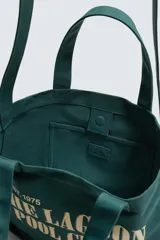 Bolso tipo shopper de algodón en color verde oscuro, con diseño de estampado tipográfico en color crema en el frente. Cuenta con dos asas de mano y cierre magnético.