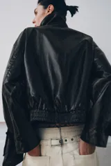 Chaqueta de efecto piel en color negro con cuello subido y bufanda desmontable integrada mediante botones. Presenta manga larga con trabillas ajustables en los puños, cierre frontal con cremallera y bolsillos laterales ocultos en las costuras. El diseño incluye un bajo con elástico en la espalda y forro interior.