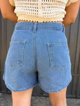 Short pollera de jean de lavado medio, con diseño cruzado asimétrico y cierre frontal con botón metálico, costuras verticales y bolsillos delanteros.