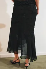 Falda midi negra de tiro medio con superposición de tejido transparente con diseño estampado y aplicación de abalorios a tono. Presenta bajo asimétrico con volantes y forro interior negro.