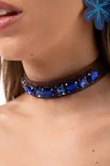 Gargantilla tipo choker de cuero sintético marrón, adornada con una hilera de cristales facetados en color azul intenso. Cuenta con cierre ajustable mediante cadena de acero quirúrgico.
