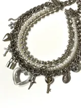 Gargantilla choker multicapa compuesta por cinco hileras que combinan cadenas de acero quirúrgico, perlas esmaltadas y perlas de hematite. Incluye múltiples dijes colgantes, destacando un corazón de cristal transparente, una llave, una cruz, un candado y una medalla religiosa.
