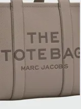 Cartera tipo tote de cuero granulado color tostado, con diseño estructurado y rectangular. Presenta doble asa superior fija y una correa larga ajustable y extraíble. En el frente, destaca el logo estampado con la inscripción 'THE TOTE BAG MARC JACOBS'.
