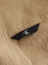 Camisa de manga larga color beige, confeccionada en tejido texturizado de algodón. Presenta cuello camisero, cierre frontal de botones y un bolsillo en el pecho con una insignia cuadrada negra con el logo 'CK' en blanco.