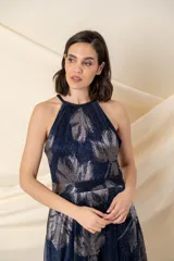 Vestido largo de fiesta en gasa plisada color azul con estampado plateado de hojas. Tiene falda amplia, cruzada y abierta, con bolsillos laterales. El escote es halter y tiene un cinto fino a la altura de la cintura.