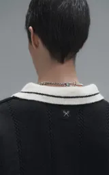 Sweater de punto negro con cuello polo en color crema, manga larga y diseño oversize. Presenta bordados en el frente con la inscripción 'Ravers', el número '90' y letras cursivas, con terminaciones en punto rib en puños y cuello.