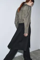 Blusa color gris con finas rayas verticales negras, cuello redondo con lazo del mismo tejido, manga larga con puño, bolsillos de parche en el pecho, bajo asimétrico y cierre frontal oculto con botones.