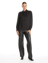 Blusa negra de acabado satinado con cuello alto fruncido, detalle de lazo para anudar y cierre lateral con botones. Presenta mangas largas estilo balloon con puños ajustados.