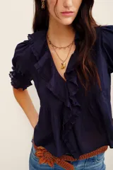 Camisa azul marino de algodón, con mangas cortas abullonadas y puños fruncidos con elástico. Presenta un amplio escote en V adornado con volados con detalles bordados y ojales a lo largo de la botonera frontal.