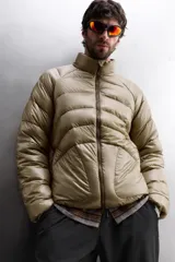 Campera puffer color beige con cuello alto, cierre frontal con cremallera y bolsillos laterales con cierre. Diseño acolchado horizontal.