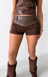 Short corto marrón con diseño combinado de cuerina lisa en la parte superior, trabillas para cinturón y detalle de tachas metálicas en la costura que une con la sección inferior de gamuza.