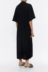 Vestido largo negro de viscosa con textura, corte recto, cuello camisero con solapas, manga corta, cierre frontal de botonadura, bolsillos plastrón en el pecho, bolsillos con tapeta en bajo delantero y cinturón de tejido.