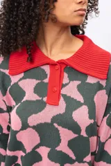 Buzo de tejido de punto con diseño estampado geométrico en tonos verde oscuro y rosado. Presenta cuello tipo polo en color rojo, cierre frontal con botones y puños acanalados en color rojo.