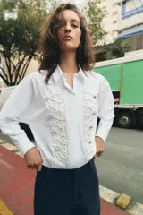 Camisa de popelín blanca con cuello solapa y manga larga. Presenta un diseño frontal con bordados en relieve y botones decorativos negros. Cierre frontal con botones ocultos.