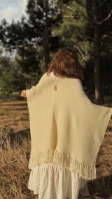 Ruana tejida en lana merino, de color crema, con diseño de punto texturizado y terminación con flecos en el ruedo. Presenta una etiqueta de cuero decorativa en la parte superior de la espalda.