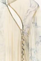 Camisa de cuello redondo y manga larga con diseño de estampado artístico en tonos azules sobre fondo beige. Presenta un cierre frontal asimétrico con botones forrados a tono y una abertura lateral en el escote.