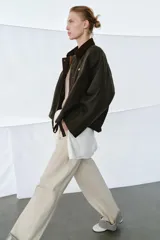 Chaqueta encerada de corte oversize, color marrón oscuro, con cuello de pana en contraste. Presenta cierre frontal con cremallera y botones a presión ocultos bajo solapa, bolsillos delanteros y mangas largas con puños ajustables.