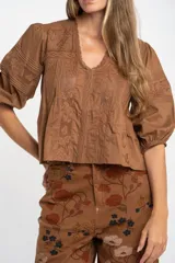Blusa de algodón color marrón con cuello en V, mangas tres cuartos con puños elásticos y detalles de encaje y alforzas en el frente.