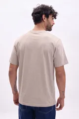 Remera oversize color beige de manga corta, con cuello redondo y un pequeño parche bordado en el pecho.