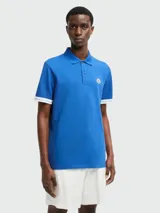 Polo de manga corta color azul, confeccionado en algodón. Presenta cuello clásico, cierre frontal con botones y detalles de franjas blancas en los bordes de las mangas. Incluye el parche con el logotipo de la marca en el pecho.