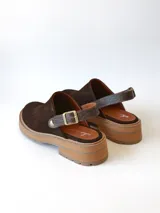 Zueco de cuero gamuzado color chocolate, con diseño de punta cerrada y tira ajustable con hebilla metálica en el talón. Presenta una suela de plataforma dentada en tono marrón claro.