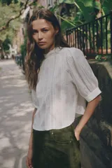 Blusa blanca de cuello alto con detalles de puntilla, manga corta abullonada y cierre posterior con botón.
