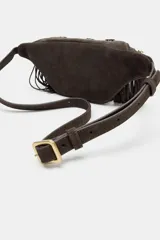 Riñonera de serraje color marrón con diseño de flecos largos en la parte frontal. Presenta detalles de tachas metálicas y ojales decorativos, cierre principal con cremallera y correa ajustable.