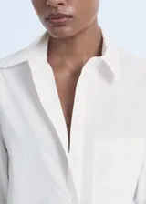 Camisa de corte recto y diseño crop, confeccionada en popelín de algodón. Presenta cuello camisero, cierre frontal con botones, mangas tres cuartos y un bolsillo en el pecho con detalle de strass.