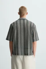 Camisa de calce relajado confeccionada en tejido jacquard con estructura abierta y diseño geométrico. Presenta cuello bowling, manga corta, aberturas laterales en el bajo y cierre frontal con botones.