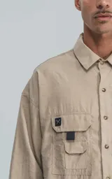Camisa de manga larga color beige, estilo técnico con calce oversize. Presenta cuello camisero, cierre frontal de botones y múltiples bolsillos de parche con solapa en el frente, algunos con tiradores negros.