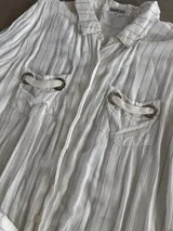 Chemise larga de rayas color blanco, con cuello camisero, manga larga y bolsillos frontales con detalles de argollas metálicas.