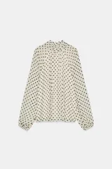 Blusa de cuello subido con lazo en la espalda, manga larga con puños elásticos, forro interior y estampado de lunares negros sobre fondo blanco.