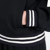 Campera deportiva New Balance modelo Track Jacket, color negro, con cierre frontal y cuello alto. Presenta puños y cintura con ribetes a rayas blancas. Lleva un pequeño logo NB bordado en el pecho.