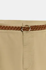 Pantalón chino color beige de tiro medio con bolsillos frontales y falsos bolsillos de vivo en la espalda. El bajo tiene un acabado con vuelta. Incluye un cinturón trenzado marrón.