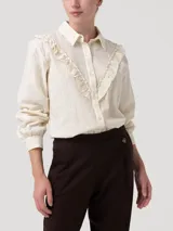 Blusa de algodón orgánico color crema con cuello camisero, cierre frontal de botones y mangas largas con puños. Presenta un diseño con volantes bordados que recorren el frente y detalles de bordado calado a lo largo de toda la prenda.