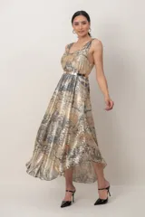 Vestido midi de fiesta color cobre con efecto metalizado y estampado abstracto en tonos grises y dorados. Cuenta con escote redondo, sin mangas, diseño cruzado en la espalda y un cinturón metálico dorado incorporado en la cintura. La falda tiene corte fluido y asimétrico, más largo en la parte trasera.