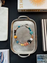 Collar corto de diseño multicolor, compuesto por cuentas grandes y medianas de diversas formas, texturas, tamaños y materiales, incluyendo cuentas de piedra, cerámica y resina en tonos turquesa, mostaza, marrón, blanco, negro y coral.