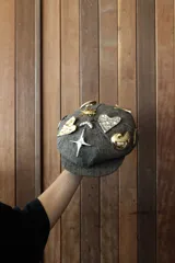 Gorra newsboy gris con diseño de espiga y apliques de pins dorados y plateados con forma de corazón, estrella, sol y otros motivos.