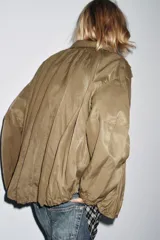 Chaqueta tipo bomber fluida color marrón, con cuello alto ajustable con cordones y stoppers, manga larga con elástico en los puños, bolsillos delanteros y bajo con pliegues. Cierre frontal con cremallera y botones a presión ocultos por solapa.