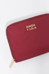 Billetera compacta de nylon color beige, con cierre de cremallera perimetral y tirador decorativo con cordón. Presenta el logo metálico de la marca en el frente.