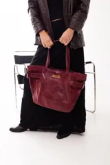 Cartera tipo tote de cuero genuino en color bordó con acabado texturizado. Presenta un diseño amplio con hebillas decorativas en los laterales, doble asa superior y una etiqueta de cuero con el logo de la marca en el frente.